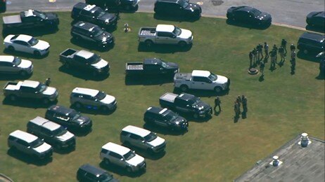 Photo shows multiple police vehicles and trucks parked in a field with law enforcement standing in the grass.